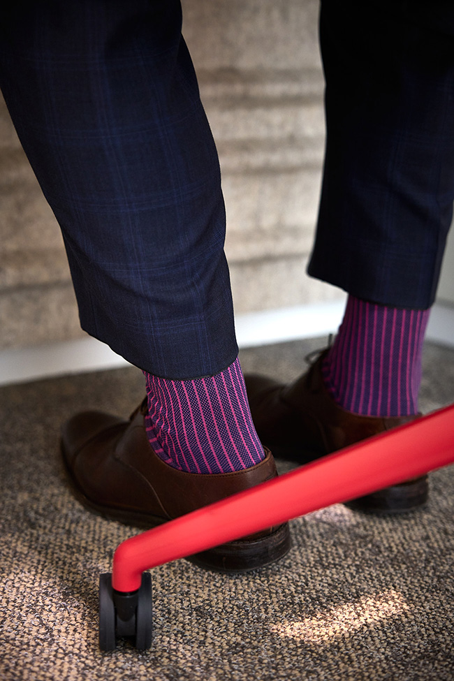 Detailaufnahme der Schuhe und bunt gestreiften Socken eines Mannes, der auf einem roten Schreibtischstuhl sitzt.